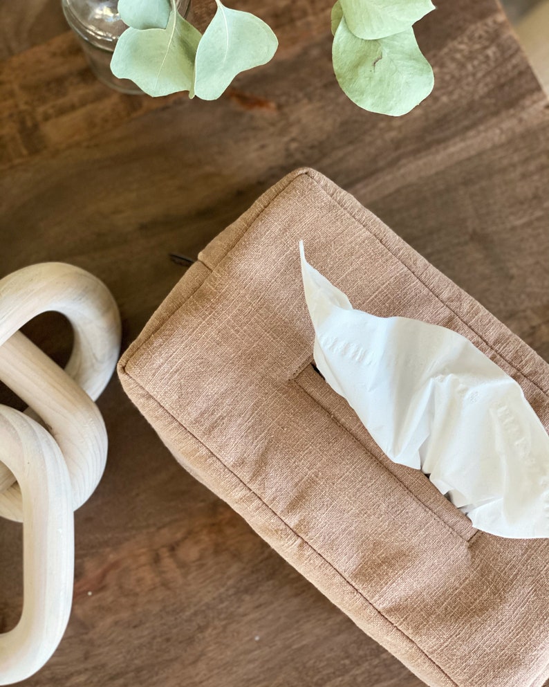 May include: A rectangular tissue box cover in a light brown fabric with a single white tissue protruding from the opening. A decorative wooden chain and sprigs of greenery are also visible.
