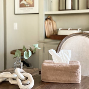 May include: A rectangular, light brown tissue box with a linen-like texture, filled with white tissues. The box sits on a dark wooden table, alongside a decorative wooden chain and a small glass vase with greenery.