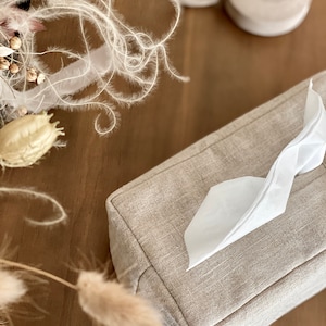 May include: A rectangular tissue box cover in a neutral linen fabric. White tissue is pulled from the top opening. A roll of white tape and dried floral elements are in the background. The tissue box cover is a light beige color.
