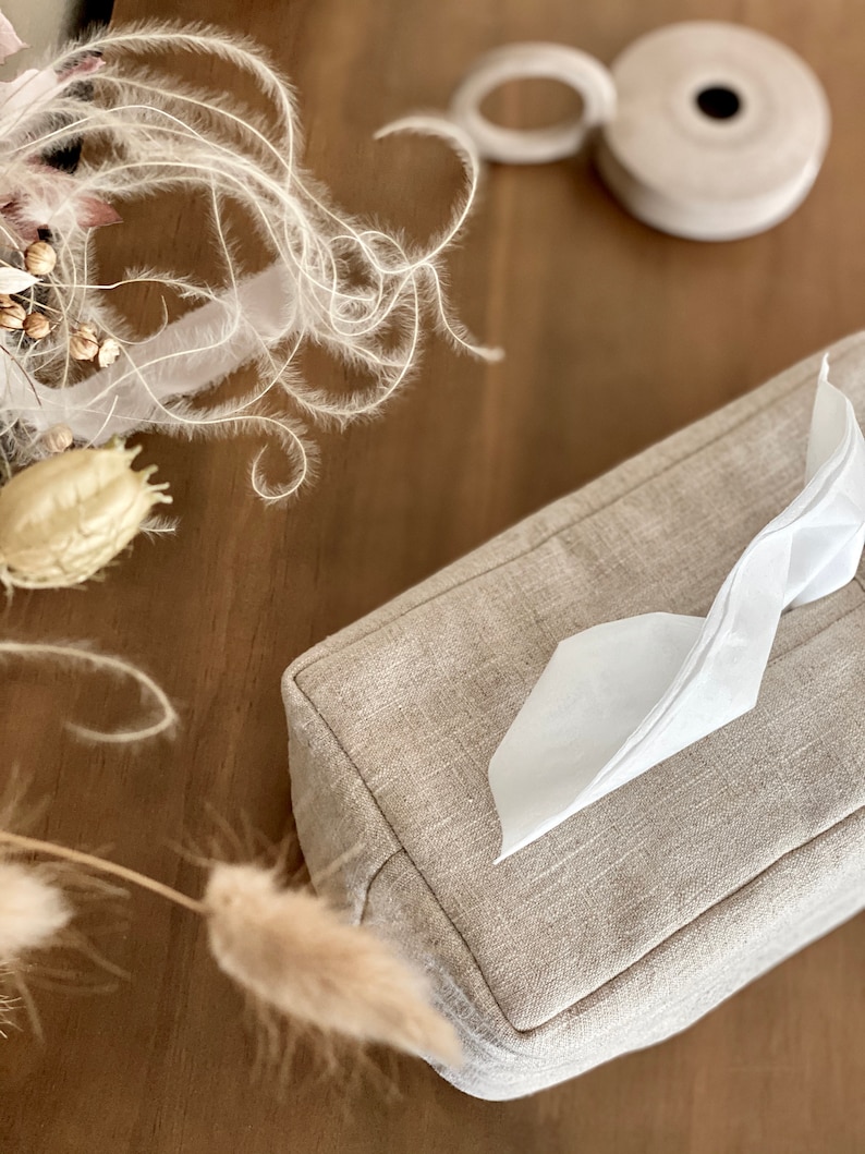 May include: A rectangular tissue box cover in a neutral linen fabric. White tissue is pulled from the top opening. A roll of white tape and dried floral elements are in the background. The tissue box cover is a light beige color.
