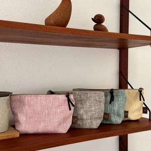 Könnte beinhalten: Vier kleine Reißverschlusstaschen in Rosa, Grau, Türkis und Gelb mit Fischgrätenmuster. Jede Tasche hat einen schwarzen Reißverschlussanhänger. Die Taschen werden auf einem Holzregal zusammen mit Holzvogelskulpturen ausgestellt.