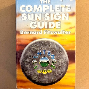 May include: A paperback book titled The Complete Sun Sign Guide by Bernard Fitzwalter. The cover features a sunset background and a circular metallic emblem displaying twelve embossed zodiac symbols surrounding a central globe graphic.