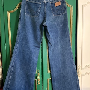 Puede incluir: Unos pantalones vaqueros azules de tiro alto con pernera ancha. La parte trasera muestra dos bolsillos con un patrón de W cosido, un parche de cuero marrón en el bolsillo derecho y un cierre central de botón en la cintura.