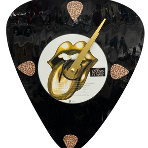 May include: A black guitar pick-shaped clock with a gold Rolling Stones tongue and lips design. The clock has four leopard print guitar picks around the edge.