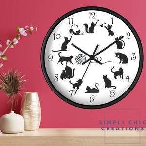 May include: A black and white wall clock with a white background and black cat silhouettes. The clock features a black frame and black hands. The numbers on the clock face are black and white. The clock is set against a pink wall.