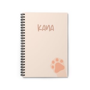May include: A cream-colored spiral notebook with the name "KANA" printed in brown text. A pink paw print is in the bottom right corner.
