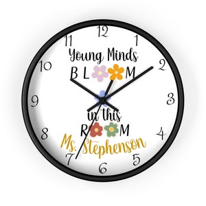 May include: A black and white clock with black hands. The clock face has the text "Young Minds Bloom in this Room Ms. Stephenson" with colorful flower illustrations.