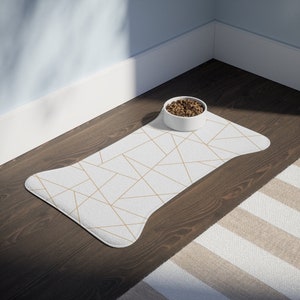 May include: White pet food mat with a geometric gold pattern. A white ceramic bowl with brown pet food is on the mat.