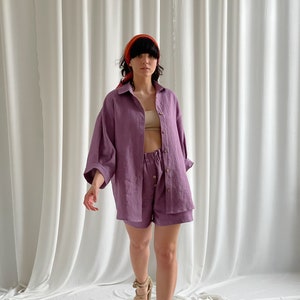 May include: A woman wearing a purple linen shirt and shorts set. The shirt has a collared neckline and long sleeves. The shorts have a drawstring waistband. The woman is wearing a red headband and sandals.