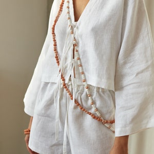 Peut inclure: Un haut en lin blanc avec un col en V et des manches longues, nou&eacute; &agrave; la taille avec un collier de perles brun et blanc.
