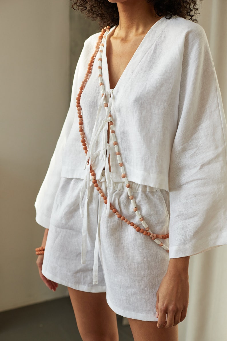 Peut inclure: Un haut en lin blanc avec un col en V et des manches longues, nou&eacute; &agrave; la taille avec un collier de perles brun et blanc.