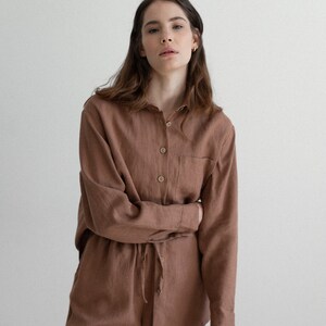 May include: A brown linen shirt and pants set. The long-sleeved shirt has a button-down front and a single chest pocket. The pants have a drawstring waist. The outfit is made of a soft, natural fabric.