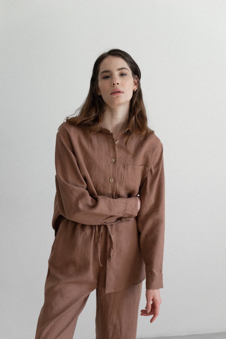 May include: A brown linen shirt and pants set. The long-sleeved shirt has a button-down front and a single chest pocket. The pants have a drawstring waist. The outfit is made of a soft, natural fabric.