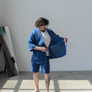 Puede incluir: Una persona vistiendo una bata de lino azul y pantalones cortos a juego. La bata está abierta y la persona está ajustando la corbata.