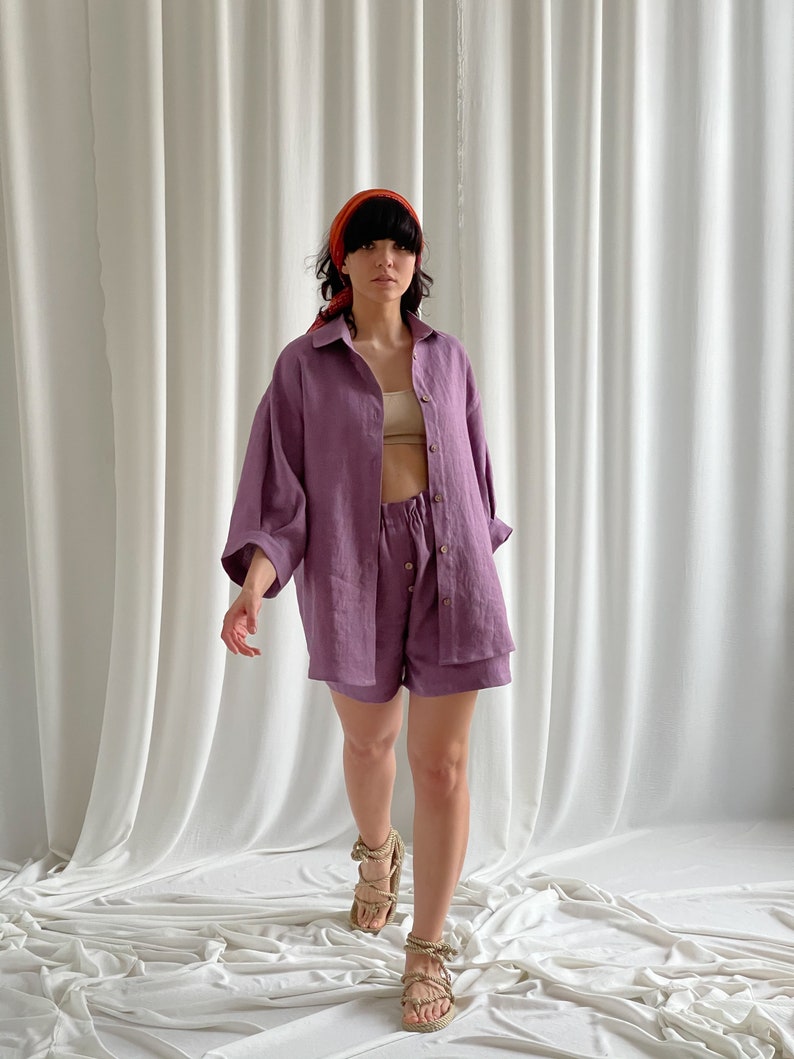 May include: A woman wearing a purple linen shirt and shorts set. The shirt has a collared neckline and long sleeves. The shorts have a drawstring waistband. The woman is wearing a red headband and sandals.