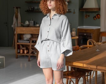 May include: A woman wearing a light gray linen shirt and shorts set. The outfit has a relaxed fit and features a button-down front and a drawstring waist. The woman is standing in a kitchen with a wooden table and chairs in the background.