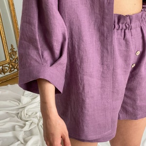 May include: A purple linen pajama set with a button-up shirt and shorts. The shirt has long sleeves and the shorts have an elastic waistband.
