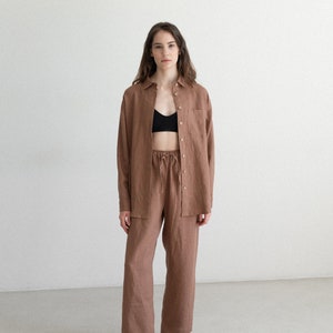 May include: A brown linen shirt and matching pants set. The long-sleeved shirt has a single chest pocket and button closures. The pants have a drawstring waist. The model is wearing a black bra and black and white sneakers.
