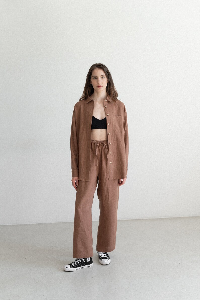 May include: A brown linen shirt and matching pants set. The long-sleeved shirt has a single chest pocket and button closures. The pants have a drawstring waist. The model is wearing a black bra and black and white sneakers.