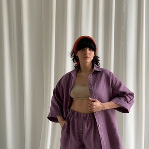 May include: A purple linen shirt and shorts set. The shirt has a button-down front and three-quarter sleeves. The shorts have an elastic waistband and a button fly.