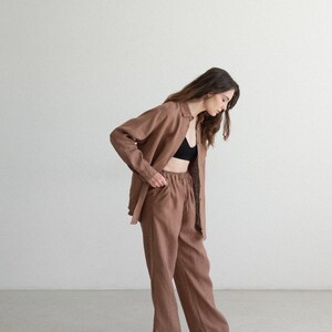 May include: A brown linen outfit consisting of a button-down shirt and matching pants. The pants have a drawstring waist. The outfit is paired with a black bra and black and white sneakers. The outfit is a neutral color.