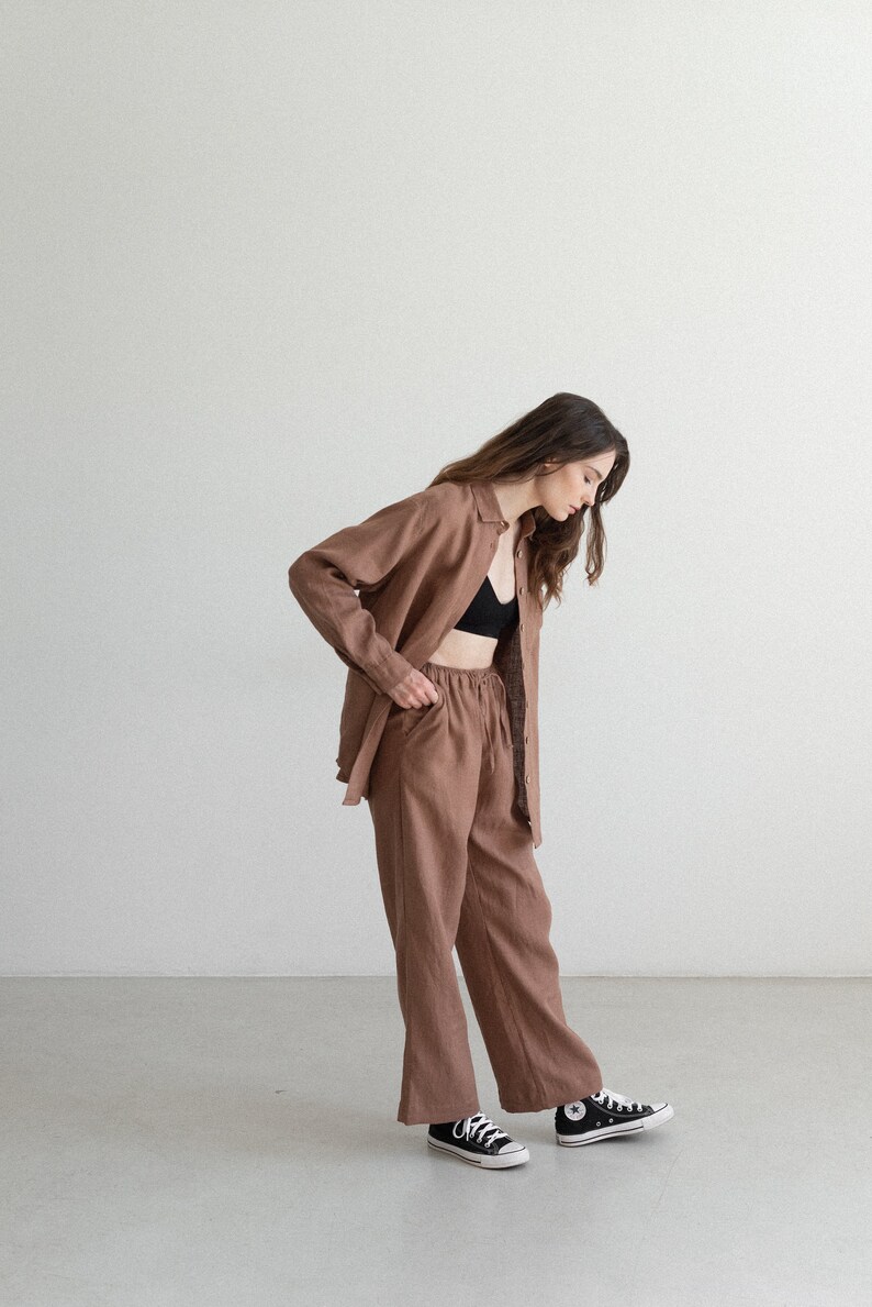 May include: A brown linen outfit consisting of a button-down shirt and matching pants. The pants have a drawstring waist. The outfit is paired with a black bra and black and white sneakers. The outfit is a neutral color.