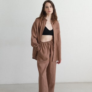 May include: A brown linen shirt and matching pants set. The shirt is button-down with a single chest pocket. The pants have a drawstring waist. The model is wearing a black bra and black and white sneakers.