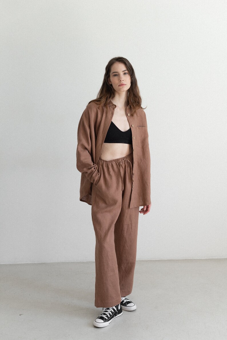 May include: A brown linen shirt and matching pants set. The shirt is button-down with a single chest pocket. The pants have a drawstring waist. The model is wearing a black bra and black and white sneakers.