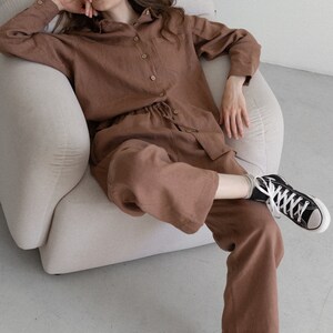 May include: A brown linen pajama set, including a button-down shirt and drawstring pants. The model is wearing black high-top sneakers with white laces. The outfit is a solid color and appears to be made of a soft, breathable fabric.