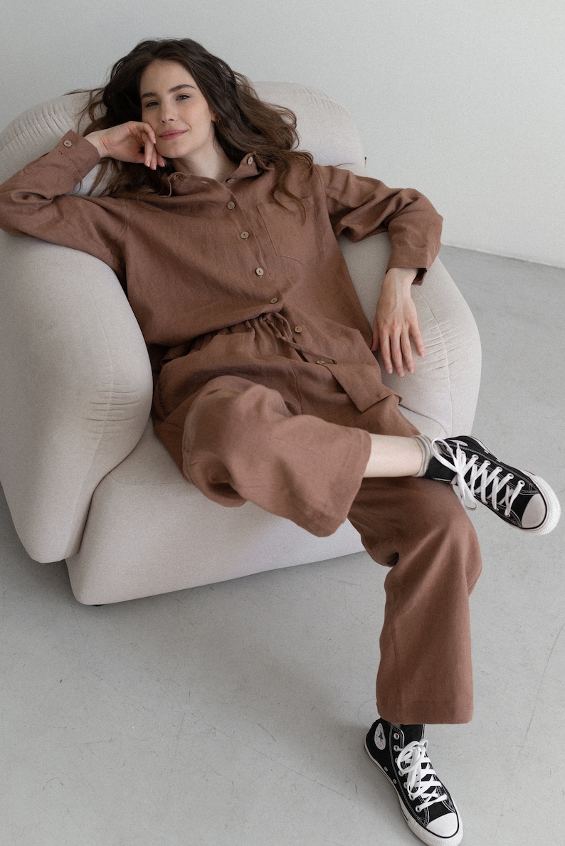 May include: A brown linen pajama set, including a button-down shirt and drawstring pants. The model is wearing black high-top sneakers with white laces. The outfit is a solid color and appears to be made of a soft, breathable fabric.