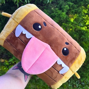 May include: A cartoon-style plush toy in the shape of a log, featuring a pink tongue, white teeth, and large black eyes. The log is brown with yellow edges and a small stick detail. The toy is designed to resemble a friendly character.