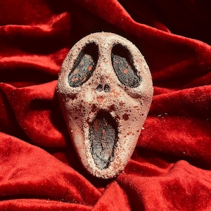 May include: A Scream mask-shaped bath bomb with a textured, speckled surface. The mask has large eye and mouth openings filled with black and red detailing. The bath bomb is set against a plush, red velvet background.