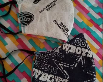 May include: Two face masks with black elastic ear loops. One mask is white with black San Antonio Spurs logos and text. The other is navy blue with white Cowboys text and star patterns. The masks are on a colorful striped background.