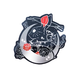 Peut inclure: Autocollant découpé représentant un lapin noir dans un croissant de lune, sur fond étoilé. Des roses rouges et des accents argentés ornent le motif. L'autocollant a une bordure argentée métallique et est sur fond blanc.