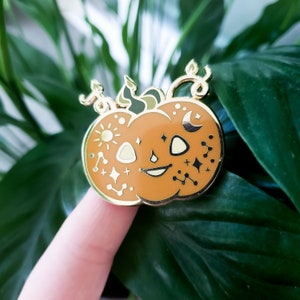 May include: A gold-colored pumpkin enamel pin with a smiling face, sun, moon, and star designs. The pin has a gold border and is set against a background of green leaves. The pumpkin is orange with cream-colored accents.