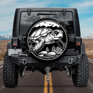May include: Black and white spare tire cover with a graphic of a sea turtle swimming in the ocean. The tire cover is on a black Jeep.