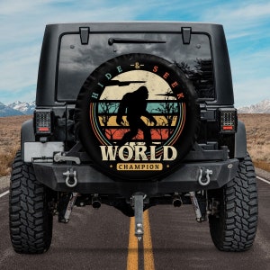 May include: A black Jeep with a spare tire cover featuring a retro-style graphic of a Bigfoot walking through a forest with the text "Hide & Seek World Champion".