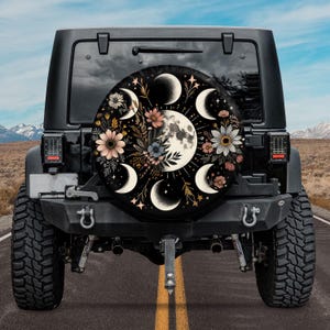 May include: A black spare tire cover with a floral design and a moon phase pattern. The cover features a full moon, crescent moons, and stars. The flowers are in shades of pink, yellow, and white.