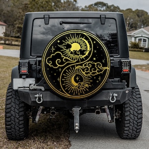 May include: Black Jeep with a spare wheel cover displaying a gold Yin Yang design with sun, moon, stars, and clouds. The circular cover is mounted on the rear of the vehicle. The Jeep is parked on a paved road.