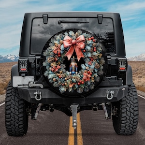 May include: A black Jeep with a spare tire cover featuring a Christmas wreath with a nutcracker figurine in the center. The wreath is decorated with red berries, pine cones, and a large red bow.