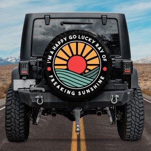 May include: A black Jeep with a spare tire cover that says "I'm a happy go lucky ray of freaking sunshine" with a graphic of a sun rising over a blue and green landscape.