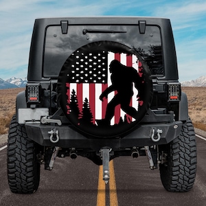 May include: A black Jeep with a spare tire cover featuring a silhouette of Bigfoot walking in front of an American flag with red and white stripes and a forest of pine trees in the background.