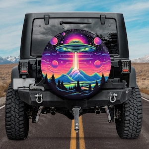 May include: A black Jeep with a colorful spare tire cover featuring a UFO flying over a mountain landscape. The cover has a pink, purple, and blue color scheme with stars and planets in the background.