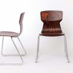 Puede incluir: Dos sillas modernas con asientos y respaldos de madera marrón oscuro. Las sillas tienen un diseño curvo y están soportadas por elegantes patas de metal plateado. Las sillas están sobre un fondo blanco liso.