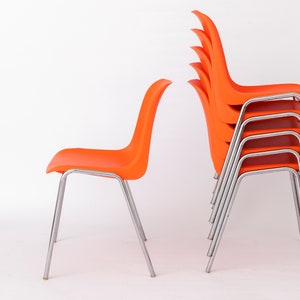 May include: A stack of six bright orange chairs with chrome legs. The chairs are stacked on top of each other, with the legs of the top chair visible. The chairs are in a white room.