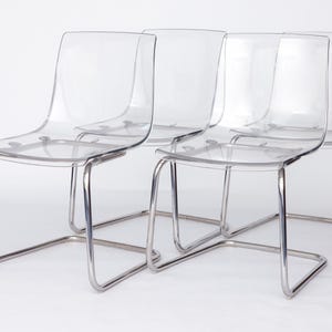 May include: Four clear acrylic chairs with chrome-colored metal frames. The chairs have a modern design with a curved back and seat. The frames are made of tubular metal. The chairs are arranged in a row.