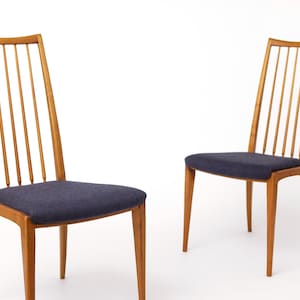May include: Two wooden dining chairs with a light brown finish. Each chair has a tall back with vertical slats and a dark blue upholstered seat. The chairs are set against a white background.