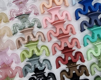 Puede incluir: Surtido de pinzas para el pelo de plástico de colores en varios diseños ondulados. Las pinzas están dispuestas en un patrón aleatorio sobre una superficie blanca.