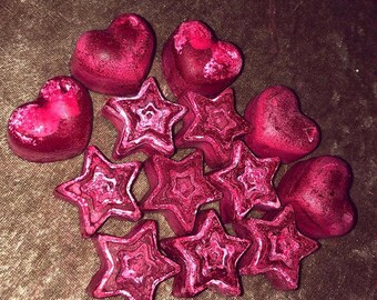 May include: A collection of pink heart and star-shaped wax melts. The wax melts are arranged in a cluster on a brown surface.