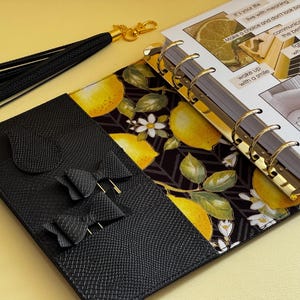 May include: A black leather planner with gold accents, featuring a lemon and floral patterned interior. Includes a black tassel and a section with bow details. The planner pages have inspirational text.